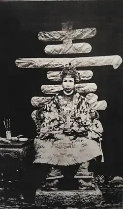 Portrait photographique d'un très jeune homme asiatique, en habit traditionnel richement décoré, assis sur un trône.