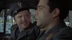 Deux hommes en uniforme filmés dans une voiture ; on aperçoit une petit Fiat à l'extérieur.