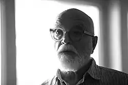 Portrait en noir et blanc d'un homme à la barbe blanche et aux lunettes rondes, dont la partie gauche du visage est dans l'ombre et la partie droite dans la lumière.