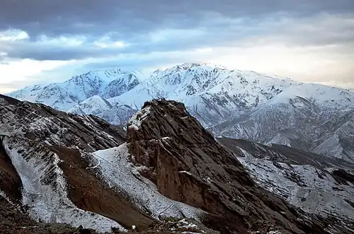 Une forteresse perchée sur un piton rocheux dans un environnement minéral et neigeux, avec de hautes montagnes en arrière-plan, ciel très couvert.