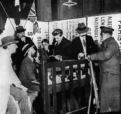 Photo en noir et blanc. Derrière une table-bureau abritée par une barrière en bois se pressent six hommes affairés en complet veston, pour la plupart chapeautés ; devant la barrière un soldat anglais reçoit un ticket de repas (il « est vu acceptant un ticket » dit la légende en anglais. La scène se passe dans un angle réduit formé de panneaux d’avis et donne une impression de confusion.