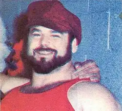 Photo d'un homme qui sourit et regarde l'appareil photo. il porte un t-shit sans manche et un béret rouge, il a une barbe noire. À sa droite, on devine un autre catcheur habillé en indien d'Amérique qui lui met la main sur son épaule.