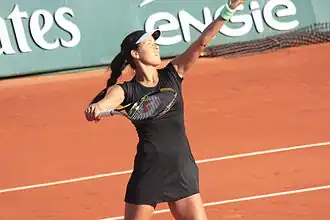 Une joueuse de tennis en robe noir lance la balle en l'air et lève sa raquette pour la frapper.
