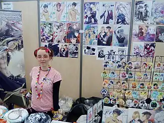 Stand de marchandises. On distingue la vendeuse et plusieurs posters ou porte-clés…