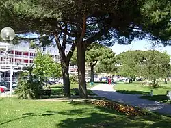 Photographie montrant des allées sablées bordées de pelouse, des massifs fleuris qui serpentent entre les pins parasols et des arbres exotiques. Au fond, les immeubles blancs et rouges du front de mer
