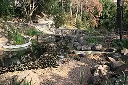 Umělé jezírko obklopené balvany. Přes jezírko vede dřevěný mostek.