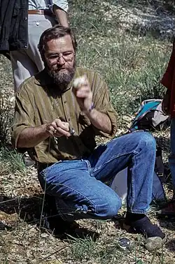 Photographie en couleur d'un homme à demi accroupi, portant une barbe et des lunettes et vêtu d'un jean et d'une chemise marron en train de reconstituer un débitage laminaire par percussion directe.