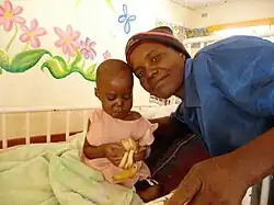 Un nourrisson pose avec un adulte dans une chambre d'hôpital aux murs décorés de peintures florales.