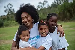 une femme avec une coiffure afro et de grandes boucles d'oreille, vêtue d'un t-shirt de l'UNICEF, tient dans ses bras trois enfants souriants. À l'arrière-plan on distingue un paysage de plaine tropicale.