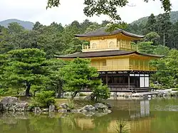 Photo couleur d'un édifice religieux carré à trois étages et aux façades dorées, se reflétant dans l'eau d'un étang, au premier plan, sur fond de forêt verdoyante.