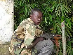 Photographie couleur d'un enfant accroupi tenant un fusil automatique.
