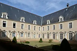 Photographie présentant un angle du jardin du cloître, dans un bâtiment à 2 niveaux de fenêtres et un toit en ardoise. Les fenêtres du rez-de-chaussée sont encadrées d'arcs surmontés d'angelots en clés de voûte. Au premier étage, sont visibles aux deux fenêtres de gauche deux mannequins en habits de religieuses.