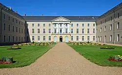 Photographie présentant un grand bâtiment en U à 3 niveaux et toit d'ardoise. Au fond, au bout d'une allée sablée encadrée de pelouses et de massifs de fleurs, l'entrée surmontée d'un fronton sculpté à colonnes.