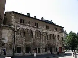 Photographie représentant un bâtiment doté de voûtes romanes, situé en ville devant une place.