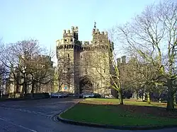 La photographie montre l'entrée d'un château, avec deux hautes tours crénelées entourant une grande porte en bois.