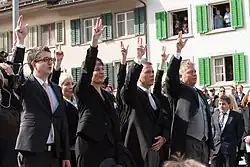 Photographie de quatre personnes, trois hommes et une femme, levant leur main droite au ciel avec le pouce, l'index et le majeur écarté.