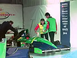 Photo du stand d'un pilote de course, où se trouvent une monoplace verte et des ingénieurs