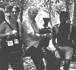 Une photographie de tournage noir et blanc où figurent trois personnes. À gauche, une jeune femme blanche en camisole avec une enregistreuse en bandoulière et une perche appuyée contre son épaule. Au centre, un homme blanc plus âgé portant des lunettes, une chemise et des pantalons avec une caméra posée sur ses genoux. À droite, un jeune homme noir en short et polo.