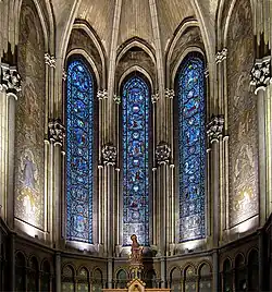 Photographie en couleurs d'une chapelle vue de face avec trois vitraux et de part et d'autre une mosaïque murale et en bas la statue de saint Jean.