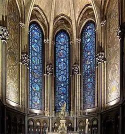 Photographie en couleurs d'une chapelle avec trois vitraux avec de part et d'autre une mosaïque murale et en bas la statue de Jeanne d'Arc.