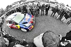 Montage photographique avec déformation sphérique et décor en noir et blanc, montrant la Citroën DS3 WRC de Sébastien Loeb entre deux rangés de spectateurs.