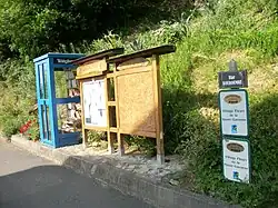 Cabine téléphonique transformée en bibliothèque de livre à échanger. Panneau d'information de la mairie et récompenses du Grand prix des années 2013 et 2014 du Village Fleuri de la Haute-Garonne