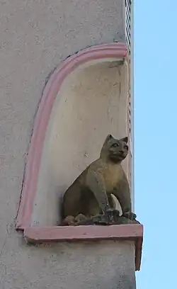 Statue, en hauteur, d'un chat assis dans une niche à l'angle d'un bâtiment. Vue selon son trois-quarts avant droit.