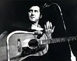 Photo en noir et blanc d'un homme muni d'une guitare derrière un micro à pied