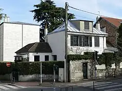 Photo en couleur d'une petite maison à un coin de rue, crépie de blanc et aux volets sombres.