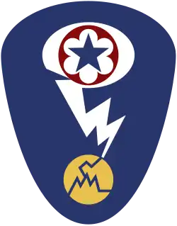 Badge d'épaule ovale avec un fond bleu foncé. Au sommet se trouve une étoile bleue entourée d'un cercle rouge qui correspond à l'écusson de l'Army Service Forces. Ce dernier est entouré d'un ovale blanc représentant un nuage en champignon. Un éclair blanc sort du nuage et frappe un cercle jaune fissuré représentant un atome.