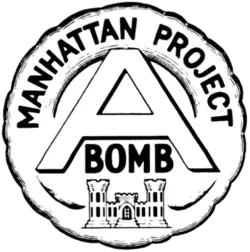 Emblème circulaire portant l'inscription « Manhattan Project » au sommet qui entoure un large « A » au centre avec le mot « bomb » écrit en dessous. L'ensemble surmonte un château qui est l'emblème du corps du génie de l'armée des États-Unis.