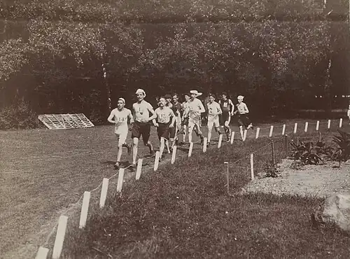 Photo en noir et blanc d'une dizaine d'hommes portants des couvre-chefs courant sur de l'herbe.
