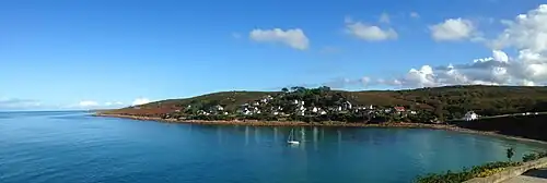 Vue d'une anse étroite, dominée par un village construit sur le flanc d'une colline prolongée par une pointe rocheuse.