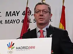 Photographie d'un homme. Il porte des lunettes, se tient debout et prononce un discours.