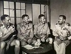 Un groupe de quatre officiers supérieurs assis ensemble en train de converser. Devant eux, une petite table sur laquelle se trouve un livre.