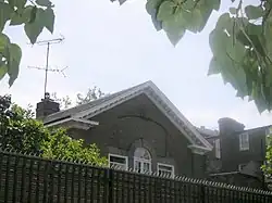 Photo sombre montrant la partie supérieure d'une maison (fenêtre arrondie, murs en briques et toit pointu), aperçue au-dessus d'un mur.