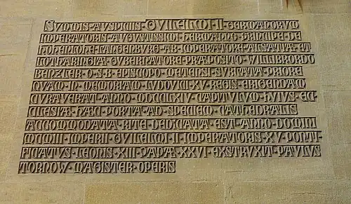 Inscription latine rappelant l'inauguration. Traduction :« Sous la plus haute souveraineté tutélaire de Guillaume II, auguste empereur des Germains, alors que le prince Hermann de Hohenlohe-Langenburg était gouverneur impérial en Alsace et Lorraine, alors que Willibrord Benzler de l'ordre de Saint Benoît était évêque de Metz, après la démolition de l'ancien portail que le chapitre de cette église a fait construire en 1764 à la mémoire du roi Louis XV, ce nouveau portail conforme au style de la cathédrale a été inauguré solennellement en l'an 1903, dans la quinzième année du règne de l'empereur Guillaume II, dans la vingt-sixième année du pontificat du pape Léon XIII. Il a été réalisé par Paul Tornow, maître d'œuvre ».