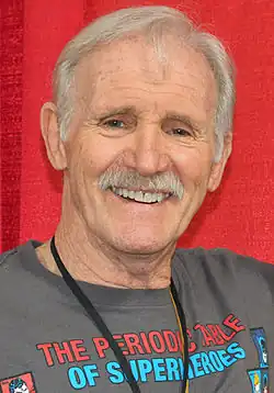 Photo en buste d'un homme âgé souriant, avec des cheveux blancs et portant un haut gris.