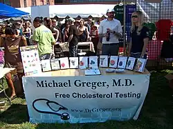 Photograph of Michael Greger at the D.C. Vegfest in 2007, standing in a white shirt in front of a booth whose banner states "Michael Greger, M.D." with "Free Cholesterol Testing" below it and his website at the bottom