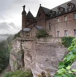 Photographie d'une falaise au sommet de laquelle est construit un bâtiment de deux étages qui possède plusieurs fenêtres et un toit pointu avec plusieurs lucarnes. Une grande colonne portant une statue de femme se trouve contre le bâtiment.