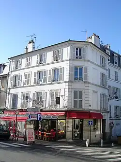 Immeuble d'angle anciens aux murs et volets blancs de quatre étage, dont un café au rez-de-chaussée.