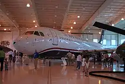 Photo prise à l'intérieur d'un hangar montrant l'avion endommagé exposé dans un musée. Des visiteurs sont présents tout autour et admirent l'avion.