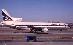 Photo couleur de l'avion, caractérisé par ses trois moteurs, dont deux sont situés sous ses ailes et le troisième sur sa queue. L'avion est peint en blanc et une fine ligne bleue sur le milieu et l'inscription Delta sur l'avant du fuselage. Il se trouve à une porte d'embarquement et des bâtiments de l'aéroport sont visibles à l'arrière-plan.