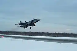 Au centre-gauche, un MiG-31, avion de chasse supersonique, décolle avec une traînée grisâtre du moteur. Au premier plan, la piste de la base aérienne, grise et striée de marques jaunes, déneigée, guide l'œil vers l'avion. En arrière-plan se trouve une ligne de taïga, et la partie supérieur de l'image est occupée par un ciel en parti nuageux, et d'un bleu froid.
