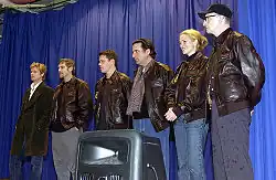 Six actors, all but one wearing a leather jacket, are photographed on a stage with a blue curtain as a backdrop.