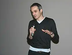 Homme de 53 ans, mince, portant un pull gris sur un T-shirt clair près du corps, courts cheveux gris, qui parle dans un micro