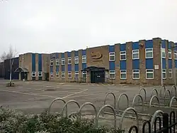 Édifice en briques brunes de forme rectangulaire avec rangées de tuiles de tôle bleues et fenêtres situé sur un stationnement pavé avec bandes jaunes. Au premier plan, supports à vélo de formes spirales.