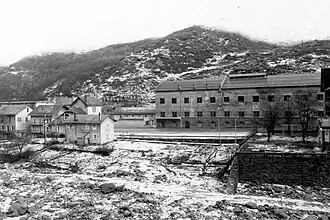 Photographie en noir et blanc d'usines proches d'une rivière, sur une carte postale en noir et blanc de l'époque ; on voit des bâtiments industriels, des logements et même des jardins, le tout dominé par une montagne enneigée.