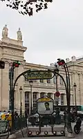 Photographie d'une bouche de métro occupant le bas de l'image, tandis que le haut de l'image montre deux allégories féminines de villes allemandes, Berlin et Cologne, surmontées de feuilles de platane se détachant sur fond de ciel blanc.