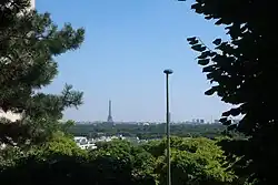 Photographie en couleur de végétation et de quelques bâtiments au premier plan et d'une tour à l'arrière-plan.